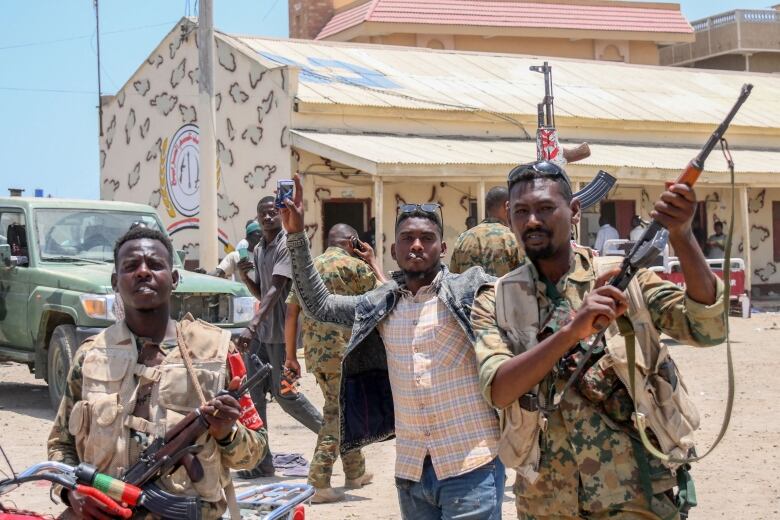 Three men hold guns and gesture toward the camera in a dirt-covered area with a vehicle and a building in the background.