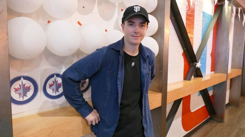 A man wearing a black shirt underneath a blue button-up leans against a wall and smiles. The wall has white balloons and Winnipeg Jets logos taped to it.