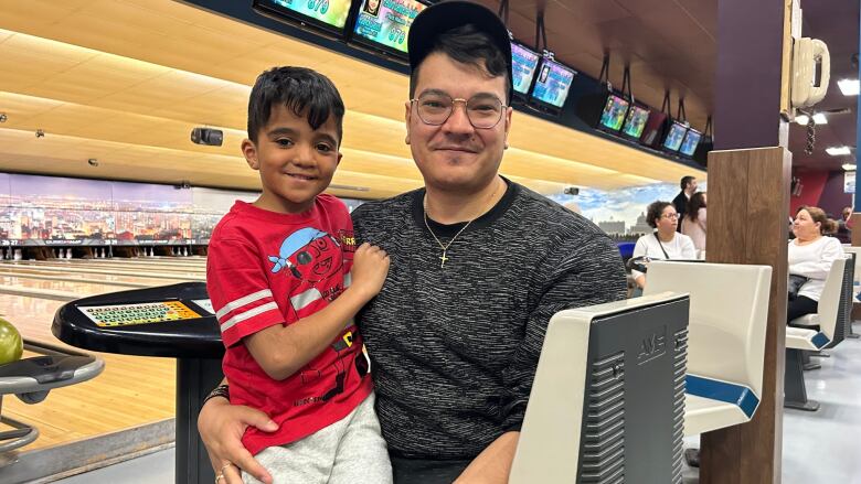 A father and son sit on a chair in a bowling alley.