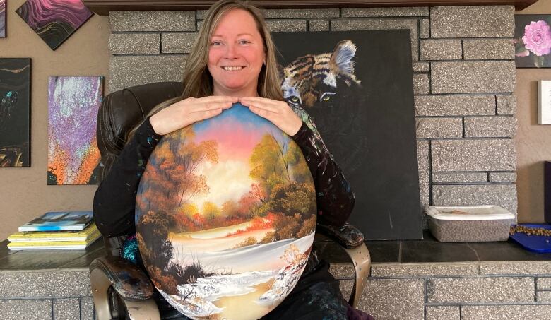 A woman poses in a home in front of a brick ledge holding up a colourful round painting of a treed landscape.