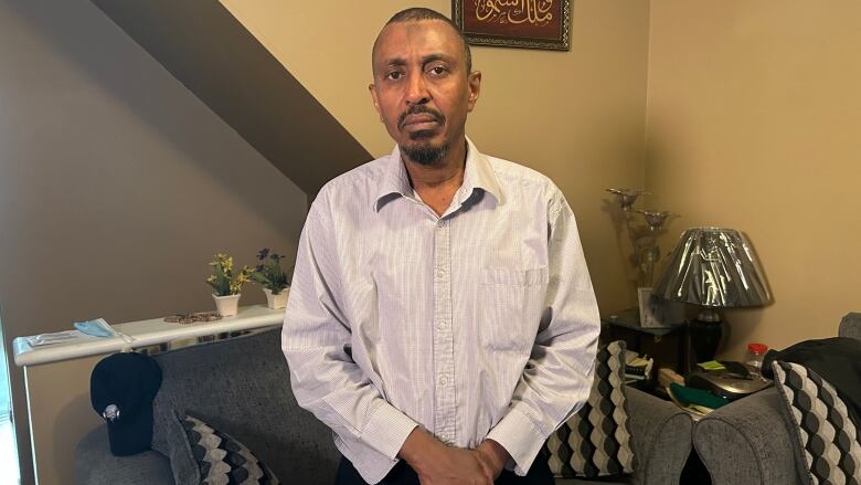 A man in a dress shirt and black pants is pictured in front of a couch and a wall with a picture frame on it.