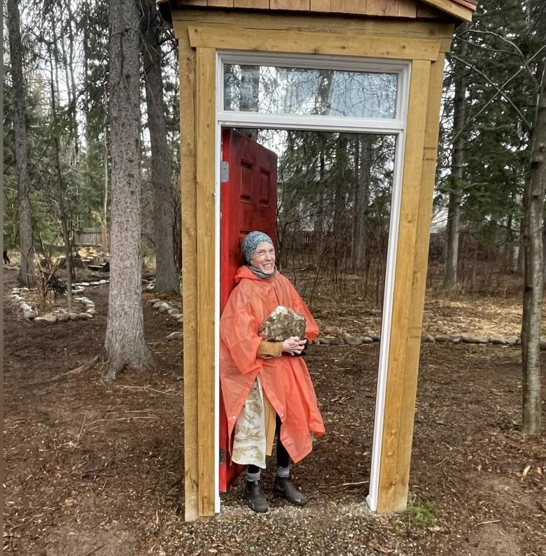 A woman with scarf is pictured holding a rock standing at a wooden door in a woodland.