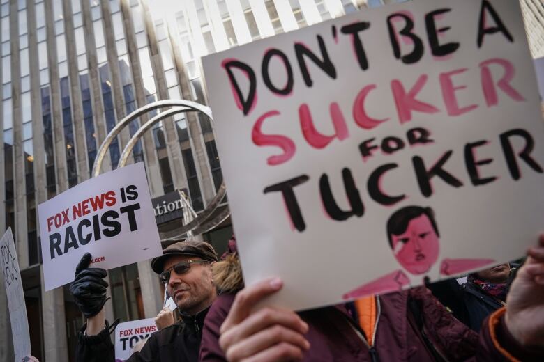 A man in the background holds a protest sign reading 