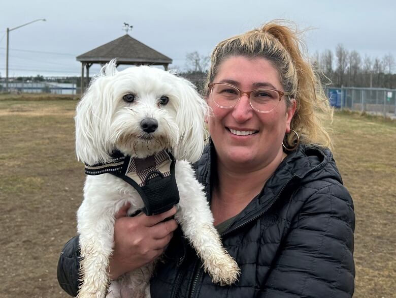 A woman, right, holding up a white small dog wearing a black harness to the left and smiling.