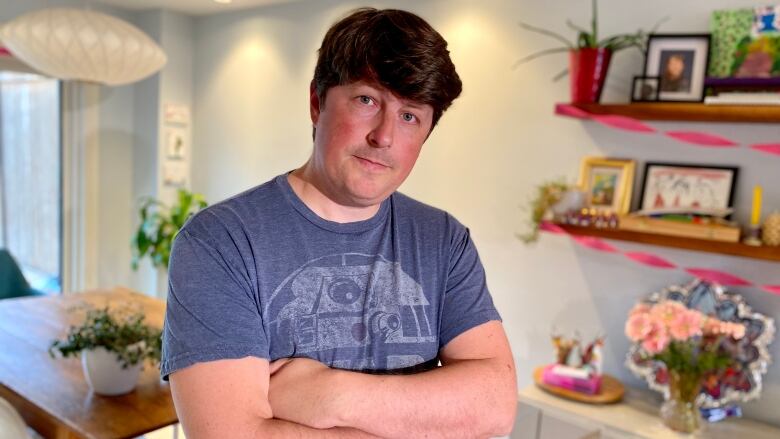 A man with short brown hair and a blue shirt is shown crossing his arms.