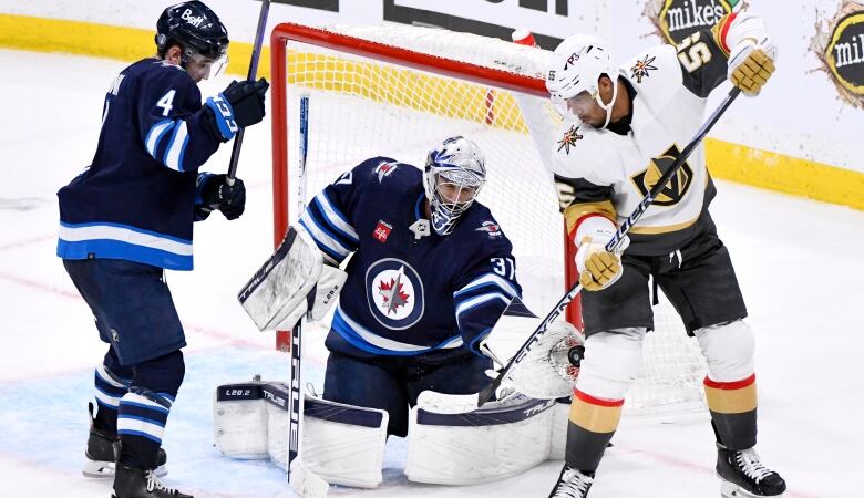 Winnipeg Jets goaltender in blue grabs the puck in his trapper with a Golden Knight player in white standing beside him with his stick raised.