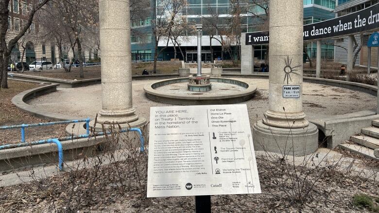 A park can be seen with tall concrete pillars, concrete steps and a central concrete fountain. The fountain is empty. 