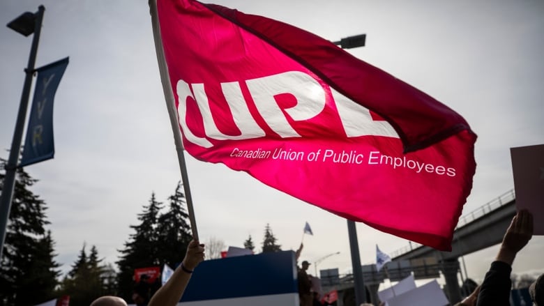 A person waves a giant red flag reading 'CUPE - Canadian Union of Public Employees.'