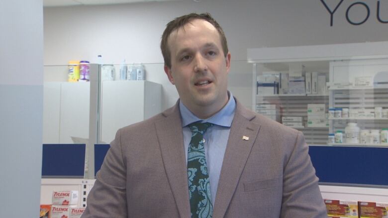 A man with short brown hair wears a grey jacket with a blue shirt and a green tie. He is standing in front of a shelf of Tylenol.