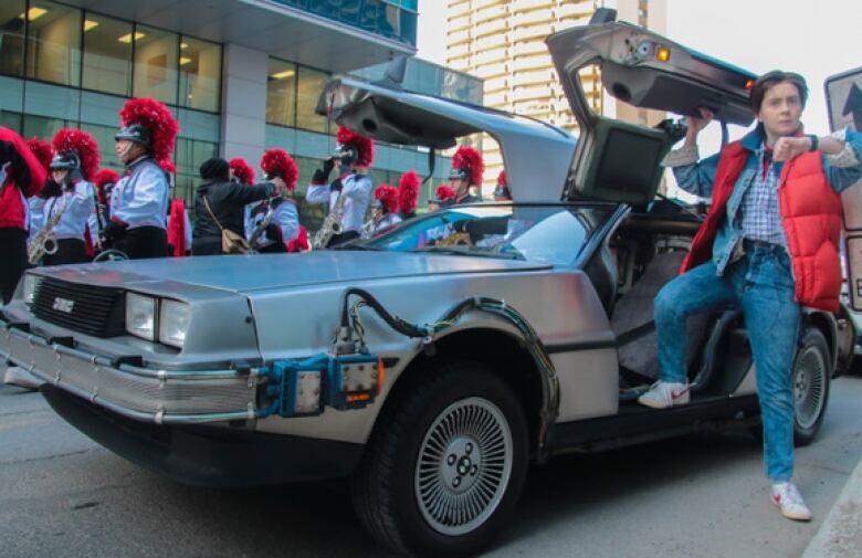 A person stands outside of a car while a marching band goes by.