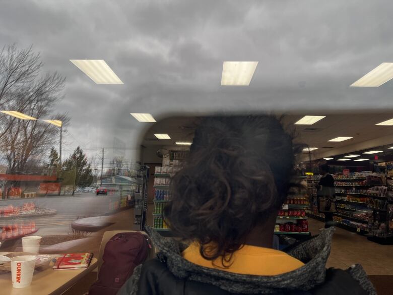 A Black woman with a curly ponytail is seen from behind through the window of a convenience store. 