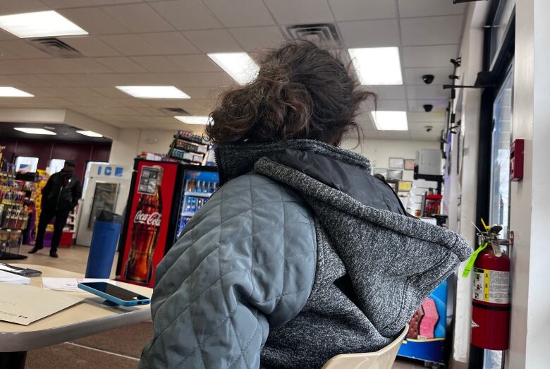 A Black woman with a curly ponytail turns her head so as not to be identified in the picture. She is inside a convenience store with papers and a cellphone scattered at the table in front of her.