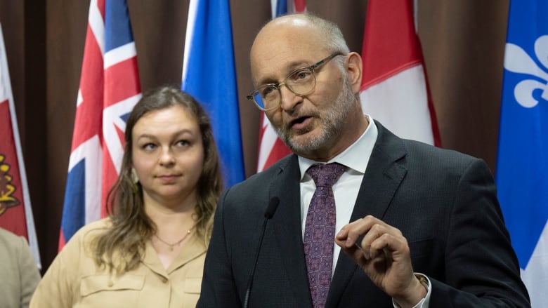 A man in a dark suit and glasses speaks at a podium. A  lady with brown hair, wearing a gold dress looks on.