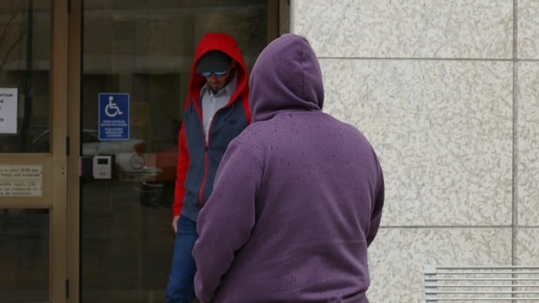 A man wearing a hat, sunglasses and hoodie is pictured with his head hanging down with a second person wearing a purple hoodie and their hood up.