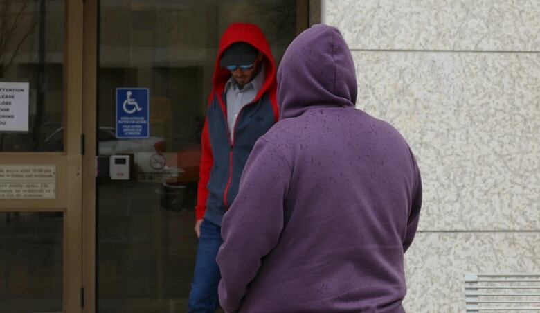 A man wearing a hat, sunglasses and hoodie is pictured with his head hanging down with a second person wearing a purple hoodie and their hood up.