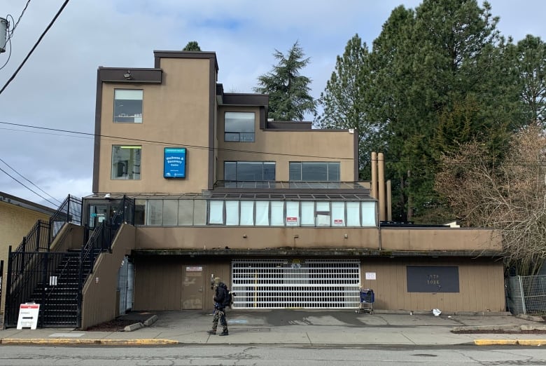 A boxy brown building with a glass room at the front, a person walks by, and smoke stacks can be seen to the right from the inhalation site. 