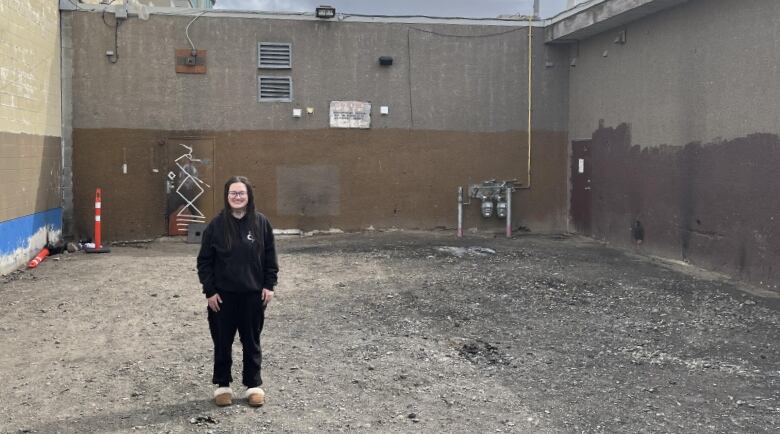 A woman in sweatpants and a sweatshirt stands in an empty dirt lot surrounded by buildings. 