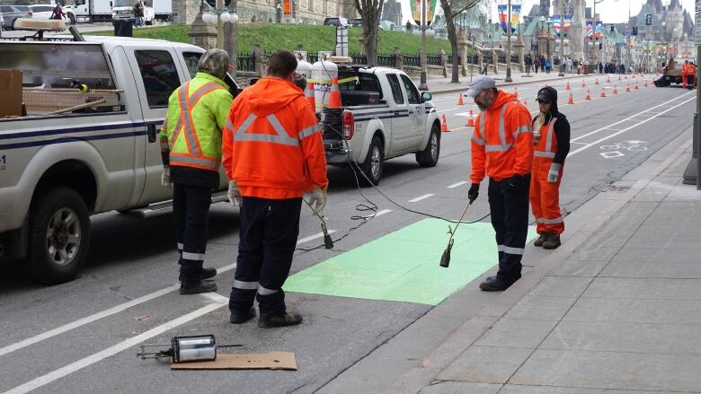 Workers in orange and yellow use a tank and hose to paint a bike lane green.