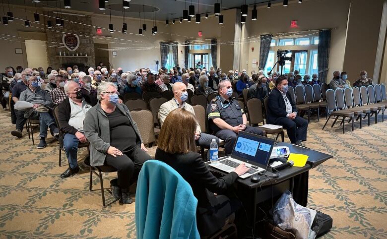 A ballroom full of people sitting in rows and wearing medical masks is shown.