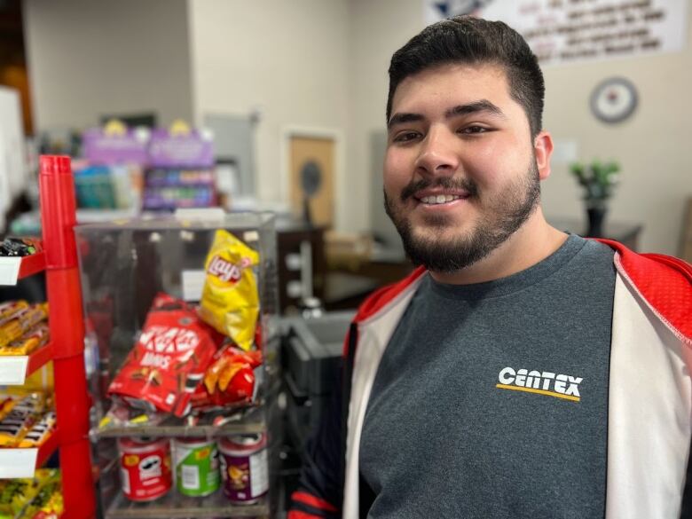 A man is shown smiling in a convenience store, with junk food behind him shown.