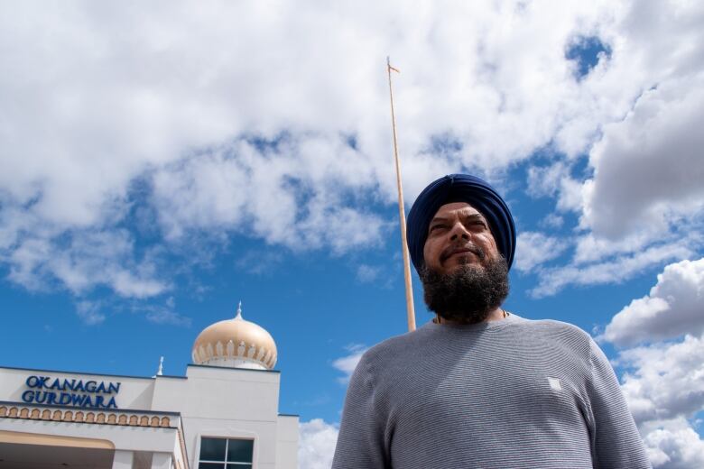 A man with blue turban and grey shirt stands in front of a flagpole next to a building with logo 'Okanagan Gurdwara.'