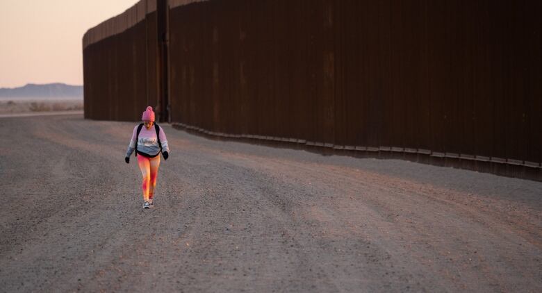 A woman walks on a dirt path near the U.S. border wall wearing runners and a toque.