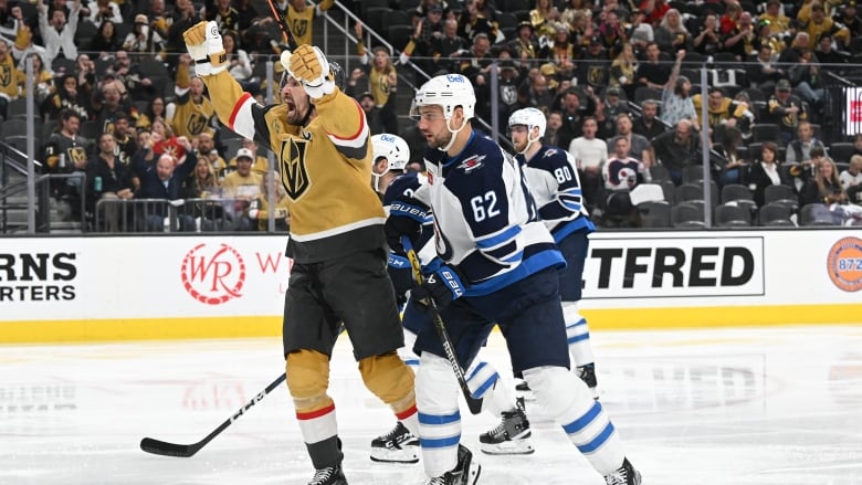 A male hockey player raises his arms while shouting in celebration as an opposing player looks on behind him and fans cheer in the stands.