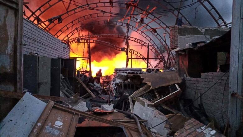 A damaged building is shown in the foreground as fire and smoke fill the sky in the background