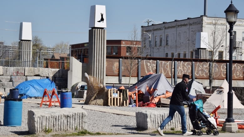 Tents are seen on a gravel lot. A man walks by on a sidewalk pushing a baby stroller. Some orange and white barricades are seen beside garbage cans.