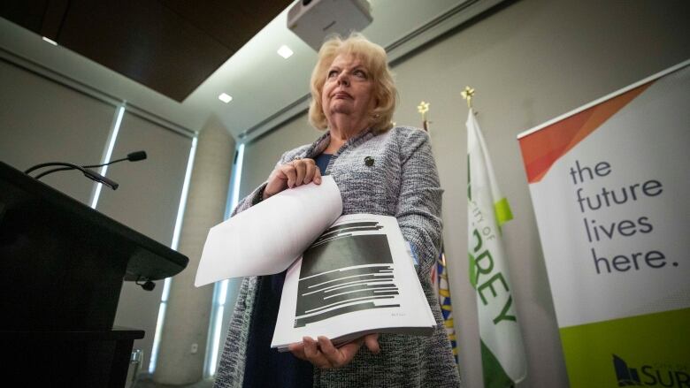 A woman with blonde shoulder-length hair standing at a podium holds open a report showing a page with blocks of blacked-out text.