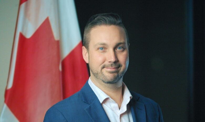 A man with dark brown hair and blue eyes wears a blue suit and stands in front of a Canadian flag.