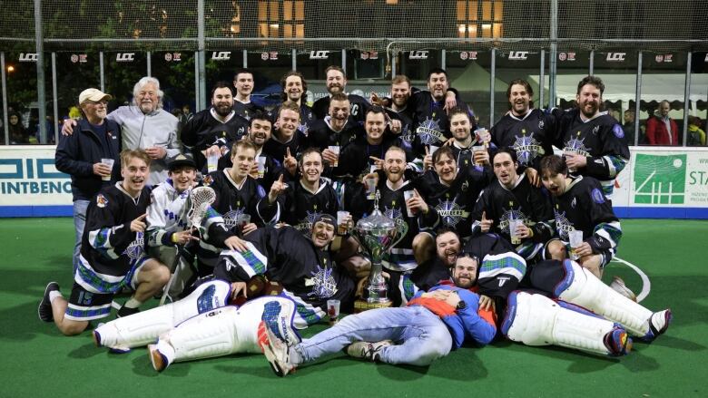 A team of lacrosse players wearing navy jerseys smile as they stand around a championship trophy.