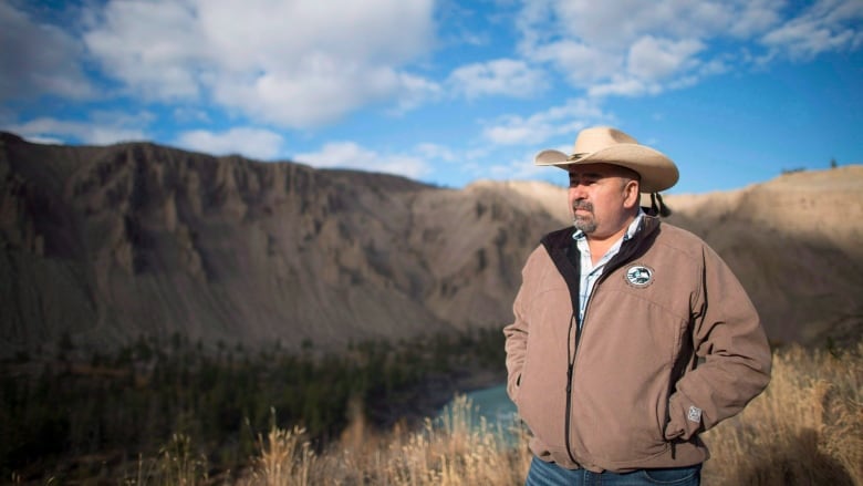 An Indigenous man wearing a cowboy hat poses in front of a hill.