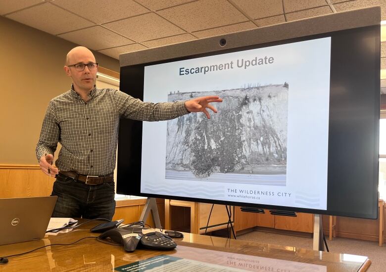 A man stands speaking in a conference room beside at a projector screen.