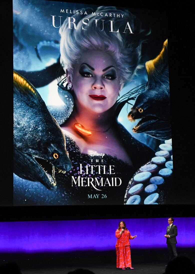 A man and a woman stand in front of a giant poster for Disney's live-action version of The Little Mermaid.