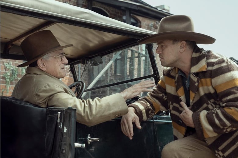 In the 1920s, an older man and a younger man speak in an early automobile.