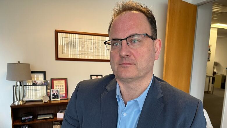 A man with brown hair and glasses wearing a blue shirt and dark blazer.