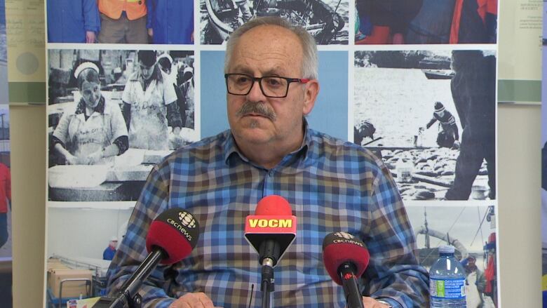 A man with glasses wearing a blue and brown flannel shirt sits in front of three microphones.