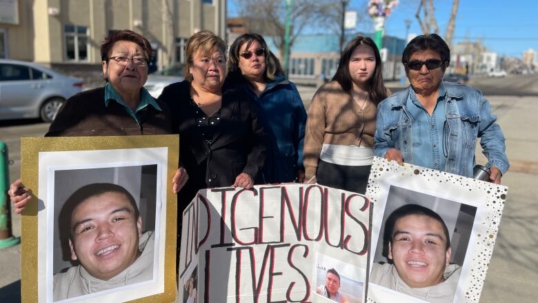 Five members of an Indigenous family stand outside a building holding pictures of a smiling man and a cardboard sign partially folded that says, Indigenous lives matter.