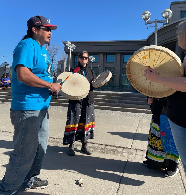 Three people bang on drums outside a building under a clear blue sky.