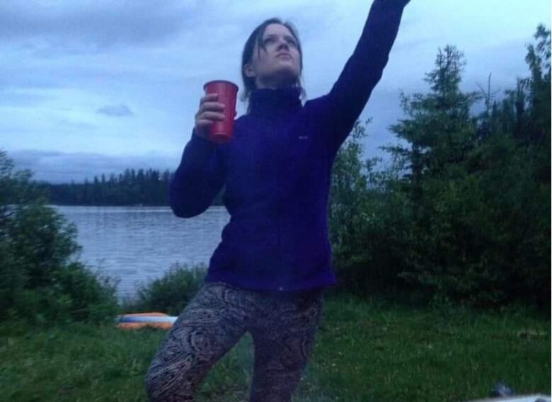 It is dusk. A woman with dark hair, pulled in a loose ponytail with bangs falling on the side of her face, is wearing a royal blue fleece jacket and paisley printed, light blue leggings, standing in front of a lake. She gazes upward, holding up a red cup in one hand while raising the other. 