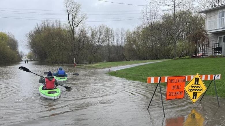 Two people paddle green kayaks down a flooded residential street.