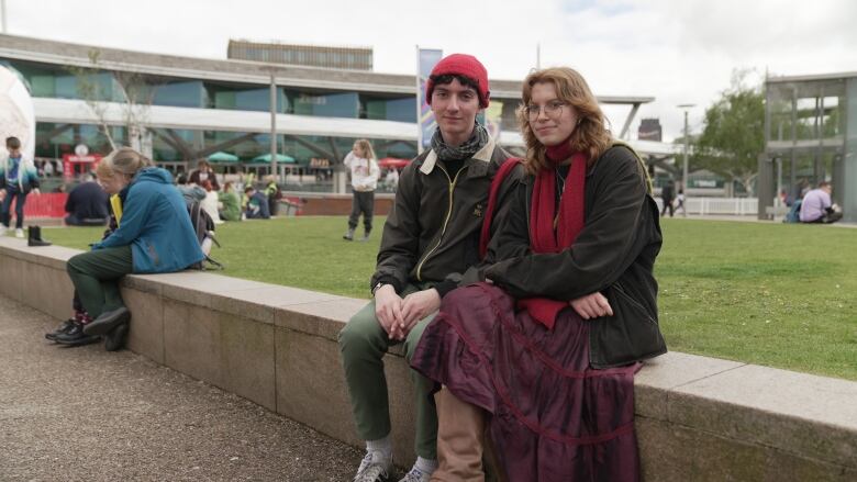 A young man and woman pose for a photo while seated next to each other on a low concrete bench that runs along the length of a parkette. Other people can be seen milling about in the background, along with a two-storey building.