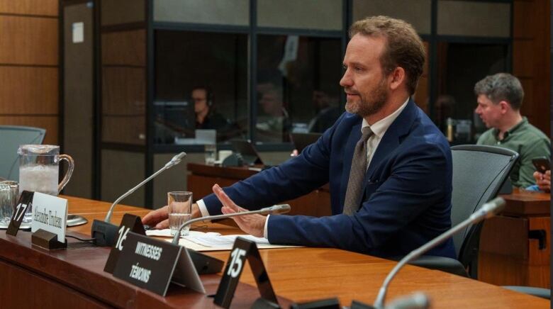 A man with a beard, wearing a blue suit, sits at a desk and responds to questions from MPs.