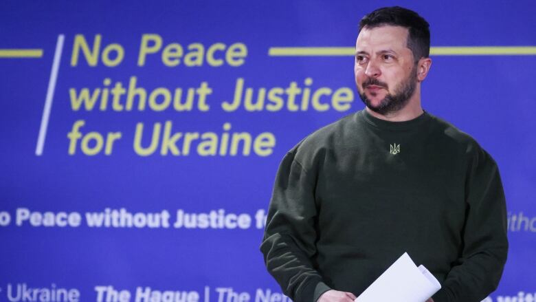 A man stands holding rolled-up paper in front of a backdrop that says 'No Peace Without Justice for Ukraine.'