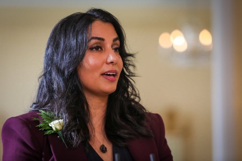 A South Asian woman wearing a mauve jacket and a white flower on her lapel speaks.