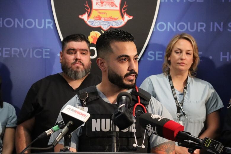 A nurse wearing scrubs and a police vest addresses a press conference with a blue background. 