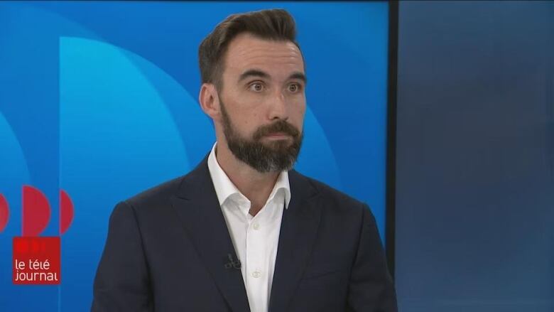 A man in a blue suit with short dark hair and beard stands in a tv studio