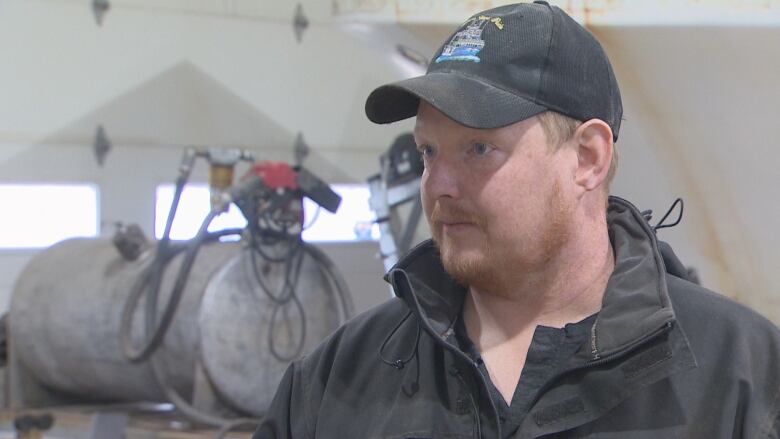 A man stands in his shed wearing a black jacket and a baseball cap.