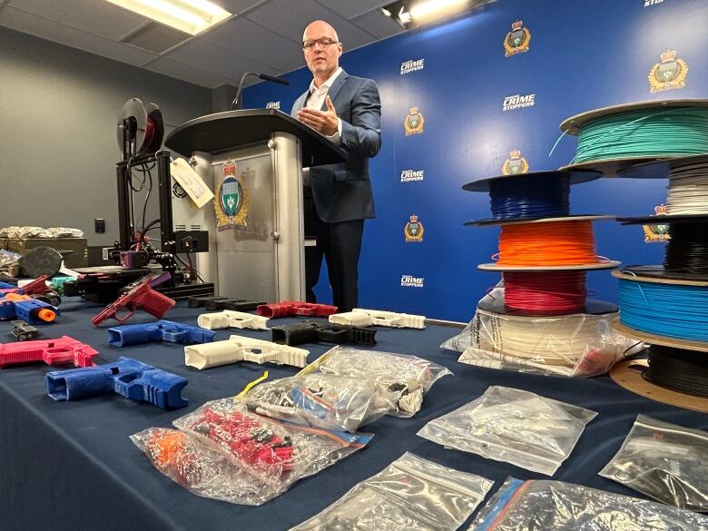 Man stands behind podium with a variety of 3D printed gun parts on a table in front of him. 